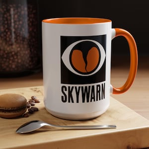 May include: White ceramic mug with an orange rim and handle. The mug features a black and orange logo with an eye and the word "SKYWARN" printed below.