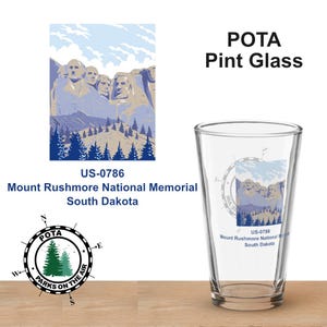 Puede incluir: Un vaso de pinta transparente con un diseño del Monte Rushmore. El vaso presenta el texto "POTA Pint Glass" y "US-0786 Mount Rushmore National Memorial South Dakota". El vaso está sobre una superficie de madera.