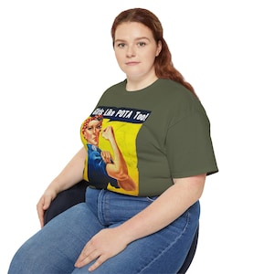 Puede incluir: Una mujer con una camiseta verde con un gráfico de estilo vintage de una mujer flexionando su bíceps y el texto "Girls Like POTA Too!" El gráfico está en un esquema de color azul, rojo y amarillo.