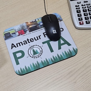 May include: A white mouse pad with a green and white logo that says "POTA Parks on the Air" and the text "Amateur Radio". The mouse pad has a green border and a design of green trees on the bottom.