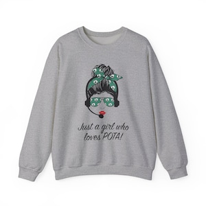Puede incluir: Sudadera gris con un gráfico de una mujer con una diadema verde y gafas de sol. El texto "Just a girl who loves POTA!" está impreso debajo del gráfico.