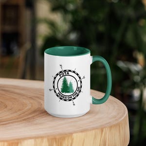 May include: White ceramic mug with a green rim and handle. The mug features a black and white compass design with the text "POTA Parks on the Air" and a green silhouette of two pine trees.