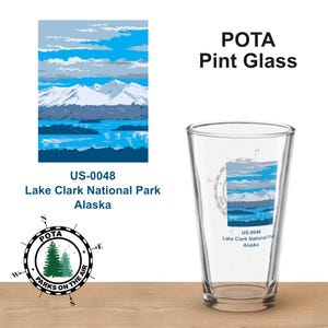 Puede incluir: Un vaso de pinta transparente con un diseño de paisaje que presenta un lago y montañas bajo un cielo azul. El vaso tiene el texto "POTA Pint Glass" y "Lake Clark National Park Alaska". También son visibles una rosa de los vientos y el texto "Parks on the Air".