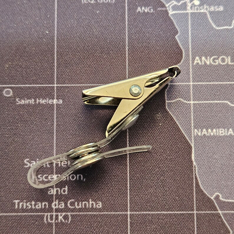 May include: A silver metal clip with a clear plastic strap. The clip is designed to hold items together.
