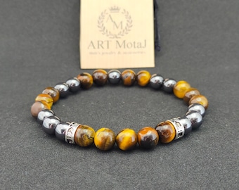 May include: A bracelet made of tiger's eye and hematite beads with silver accents. The bracelet has a silver charm with the text "ART Motaj".