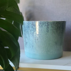 May include: A light blue ceramic planter with a textured, ombre finish. The planter has a light gray color at the top that fades to a lighter blue at the bottom.