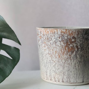 May include: A white ceramic planter with a distressed, crackled finish and a subtle copper glaze. The planter has a round shape and a smooth, slightly textured surface.
