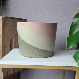 May include: A concrete planter with a pink, beige, and green color scheme. The planter is cylindrical and has a smooth, slightly textured surface.
