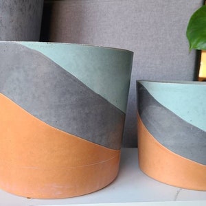 May include: Two round concrete planters with a geometric design. The larger planter is orange, gray, and light blue. The smaller planter is the same design but in a smaller size.