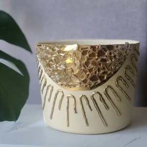 May include: A white ceramic planter with gold accents. The planter has a geometric design with a gold mosaic pattern and gold drips around the rim.