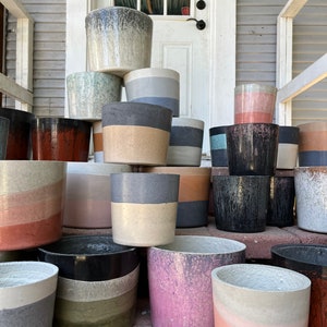 May include: A collection of 15 different concrete planters in various sizes and colors, including gray, pink, brown, and green. The planters are stacked and arranged on a wooden porch.