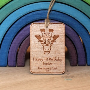 May include: A wooden tag with a giraffe illustration and the text "Happy 1st Birthday Jessica Love Mum & Dad" in front of a rainbow-coloured wooden toy.