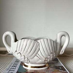 May include: White ceramic swan-shaped bowl with gold accents. The bowl has a scalloped edge and a raised base. The swan's necks form the handles of the bowl.