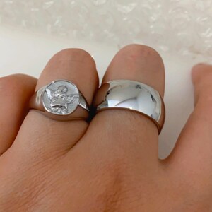 May include: Two silver rings, one with a raised design of an angel with wings, and the other a plain band.