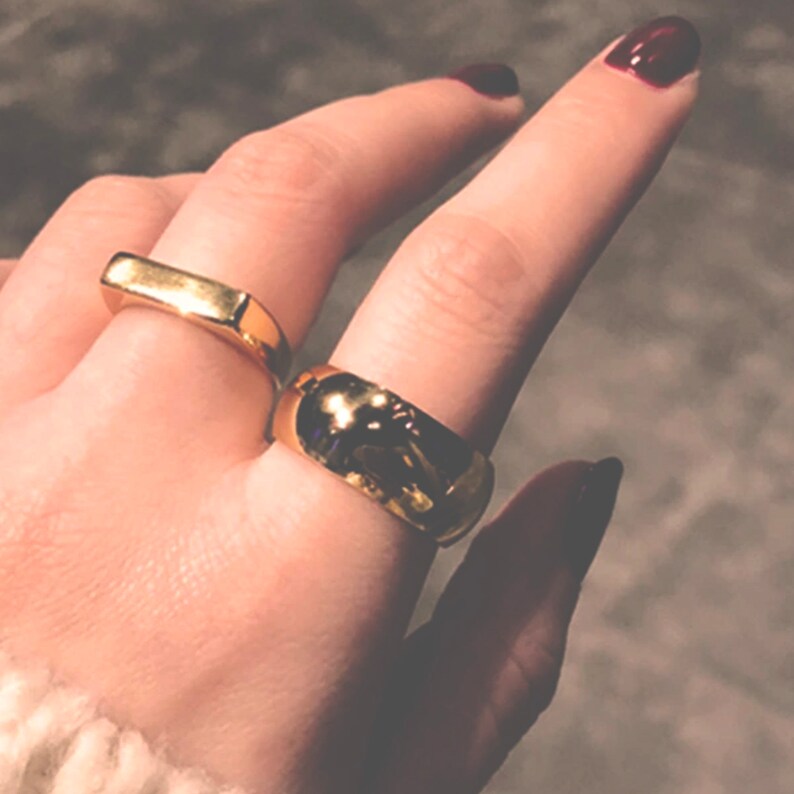 May include: Two gold rings on a hand. The ring on the index finger is a thick, rounded band. The ring on the middle finger is a flat, rectangular band.