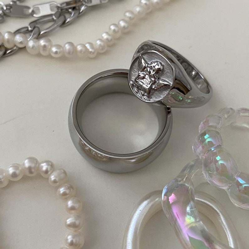 May include: Two silver rings, one with a raised angel design, on a white surface with pearl necklaces and iridescent plastic rings.