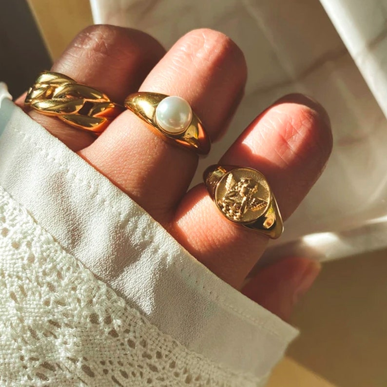 May include: Three gold rings worn on a hand. The ring on the index finger has a large pearl. The ring on the middle finger has a design of an angel with wings. The ring on the pinky finger is a thick chain link design.
