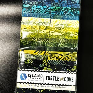 May include: A package of 40 fabric strips in various shades of blue, green, and yellow. The strips are made of batik fabric and feature a variety of patterns. The package is labeled "Island Batik Turtle Cove" and includes the text "The Best From Bali!" and "2 1/2" Strips 40 Pieces 20 Fabric Designs".