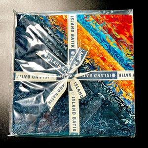 May include: A square package of batik fabric squares in shades of blue, orange, and red, tied with a ribbon that reads "ISLAND BATIK". The fabric has a variety of patterns and textures, and the package is wrapped in clear plastic.