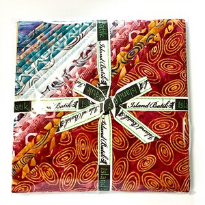 May include: A fabric bundle featuring various batik fabric patterns in shades of blue, teal, red, and yellow. The bundle is tied with a green and white ribbon that reads "Island Batik". The fabric is packaged in clear plastic.