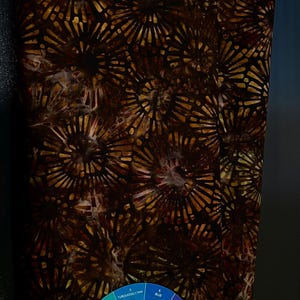 May include: A piece of fabric featuring a repeating pattern of dark brown and black circular, sunburst-like motifs with radiating lines. A circular color wheel with numbered segments from 1 to 12 is placed at the bottom center of the fabric.