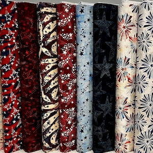 May include: Eight vertical bolts of fabric featuring patriotic themes. Patterns include red, white, and blue star motifs, swirling firework designs, and abstract starbursts on cream, red, navy blue, and light blue backgrounds in various textures.