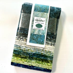 May include: A bundle of fabric squares in shades of blue, green, and gray, wrapped in clear plastic. The label reads "Shoreline 40 Karat Jewels". The fabric has a marbled, watercolor-like appearance, evoking a coastal theme. The fabric is likely for quilting or crafting.