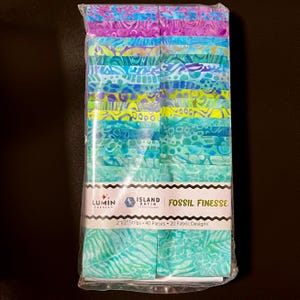 May include: A package of fabric strips in shades of blue, green, purple, and yellow. The fabric has a batik design. The package is labeled "Fossil Finesse" and contains 2 1/2" strips. The package contains 40 pieces and 20 fabric designs.