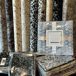 May include: Assortment of fabric rolls and pre-cut squares in various patterns and colors. The rolls display geometric, floral, and abstract designs in shades of gray, beige, and black. A package of pre-cut squares is labeled "Echoes In Stone".