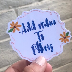 May include: White sticker with a pink outline and two orange flowers. The sticker says "Add value to others" in blue text.