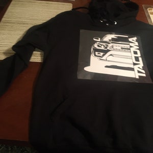 May include: Black hooded sweatshirt with a white graphic of a truck and the word "TACOMA" in white. The sweatshirt has a front pocket and a drawstring hood. The image is centered on the front of the sweatshirt.