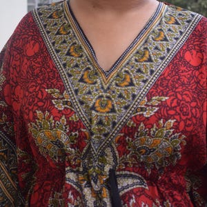 Op de afbeelding: Een rode kaftan met een V-hals en een gedetailleerd bloemen- en geometrisch patroon in geel, groen en zwart. De stof heeft een vloeiend ontwerp, geschikt voor warm weer. De halslijn is afgewerkt met een contrasterende rand.