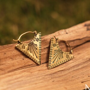 May include: A pair of gold-toned hoop earrings featuring a triangular pendant design. Each pendant is engraved with a sunburst pattern, small geometric squares, and a series of dots along the edges. The earrings include a thin wire hoop closure.
