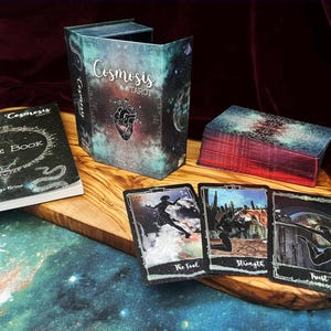 May include: A Cosmosis Tarot deck set featuring a guide book, a storage box with a heart illustration, and a deck of cards. Three cards are displayed face-up, showing The Fool, Strength, and Trust, each with dark, celestial-themed artwork.
