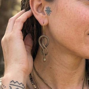 May include: A bronze-toned earring featuring a coiled snake design with a small, clear stone accent at the bottom. The earring is suspended from a thin wire hook and hangs vertically, showcasing a textured, metallic finish and a slender, curved shape.