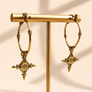 May include: A pair of gold-toned hoop earrings featuring a textured finish and a dangling diamond-shaped charm. Each charm displays a central floral pattern with small etched dots and pointed accents at the four corners, suspended from a thin wire hoop.