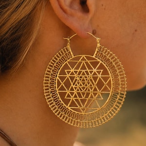 May include: A large, circular gold hoop earring featuring an intricate, geometric Sri Yantra pattern in the center. The outer edge of the hoop is decorated with a delicate, wire-like filigree design. The earring is secured with a thin metal wire clasp.