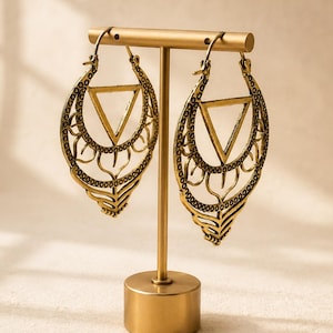 May include: A pair of antique gold-toned hoop earrings featuring an intricate filigree design with a central triangle cutout. The earrings are suspended from a T-shaped metal display stand with a circular base, showcasing the detailed metalwork.