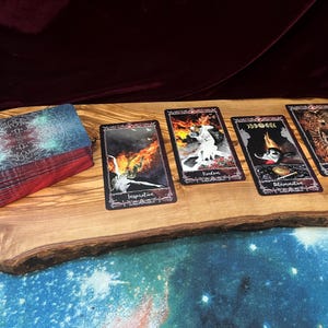 May include: A deck of tarot cards with a dark, intricate back design and four face-up cards labeled Inspiration, Freedom, Determination, and Dance. The cards feature artistic illustrations of figures and fire, arranged on a natural wood slab surface.