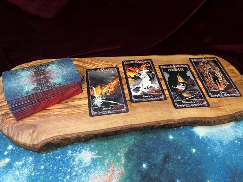 May include: A deck of tarot cards with a dark, intricate back design and four face-up cards labeled Inspiration, Freedom, Determination, and Dance. The cards feature artistic illustrations of figures and fire, arranged on a natural wood slab surface.