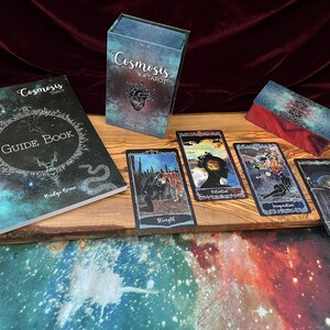 May include: A Cosmosis Tarot deck set including a guidebook, a rectangular storage box, and four tarot cards featuring celestial and surreal illustrations. The cards display the titles Strength, Potential, Inspiration, and Lust in a dark, cosmic color palette.