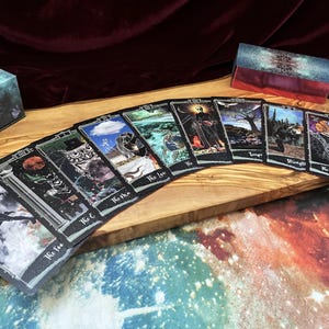 May include: A deck of tarot cards spread out in a fan shape, featuring dark, celestial, and nature-themed illustrations with white borders. The cards include titles like The Fool, The Magician, The Lovers, and The High Priestess, alongside a matching box.