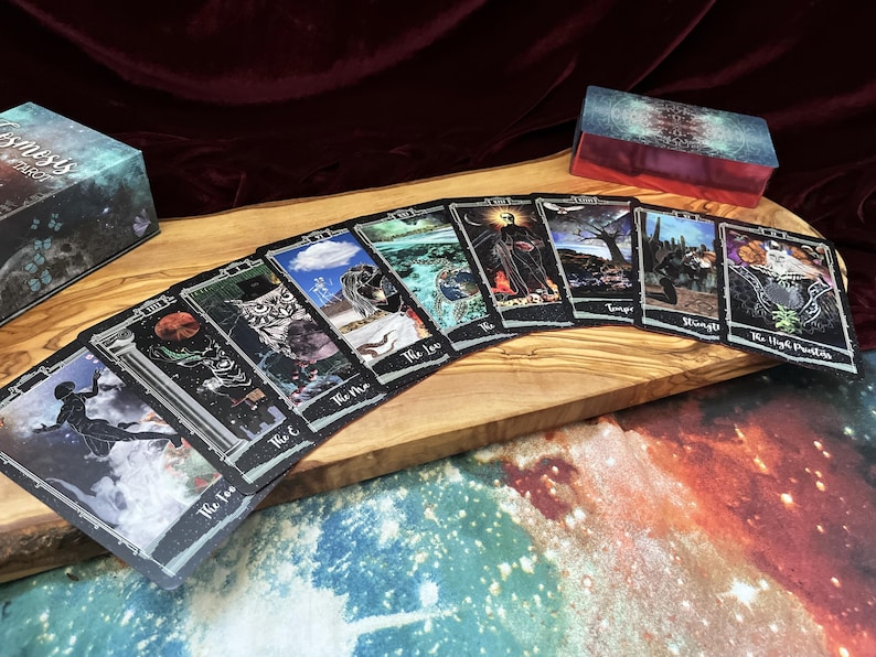 May include: A deck of tarot cards spread out in a fan shape, featuring dark, celestial, and nature-themed illustrations with white borders. The cards include titles like The Fool, The Magician, The Lovers, and The High Priestess, alongside a matching box.
