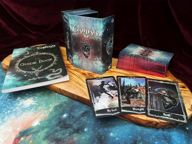 May include: A Cosmosis Tarot deck set featuring a guide book, a storage box with a heart illustration, and a deck of cards. Three cards are displayed face-up, showing The Fool, Strength, and Trust, each with dark, celestial-themed artwork.