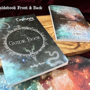 May include: Two paperback guidebooks for the Cosmosis Tarot deck, showing front and back covers. The covers feature a dark space-themed background with nebula colors, a central circular emblem, a deer head, a snake, and the text Guide Book.