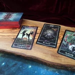 May include: A deck of tarot cards with red-edged sides and three face-up cards labeled Confrontation, The Hermit, and Maturity. The cards feature dark, detailed illustrations and are arranged on a light-colored wooden board with a natural edge.