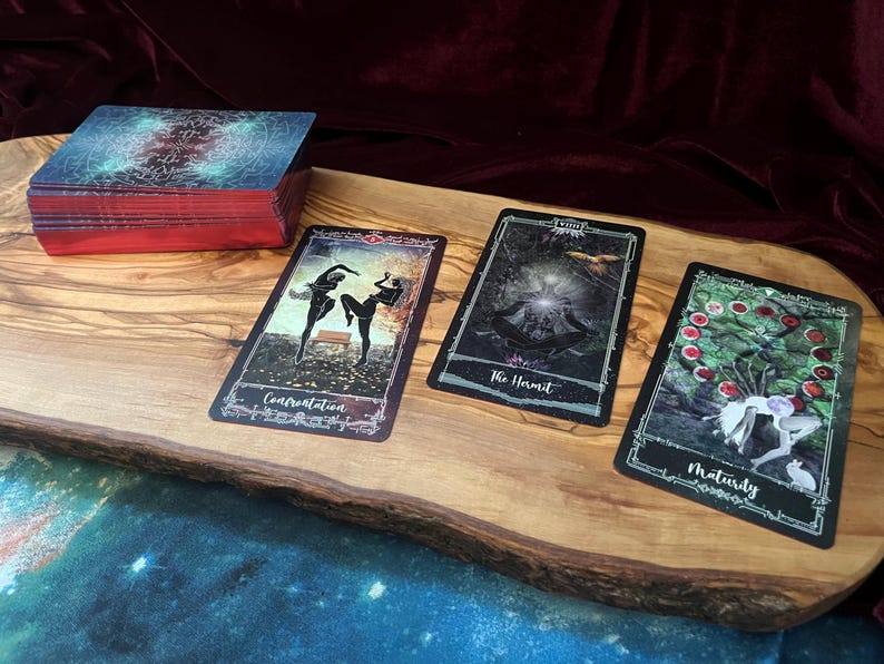 May include: A deck of tarot cards with red-edged sides and three face-up cards labeled Confrontation, The Hermit, and Maturity. The cards feature dark, detailed illustrations and are arranged on a light-colored wooden board with a natural edge.