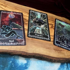May include: Three rectangular tarot cards with black borders and intricate white filigree designs. The left card shows two figures embracing under a planet with the text Bond. The center card depicts a figure in a forest with the text Perspective. The right card shows three figures with the text Celebration.