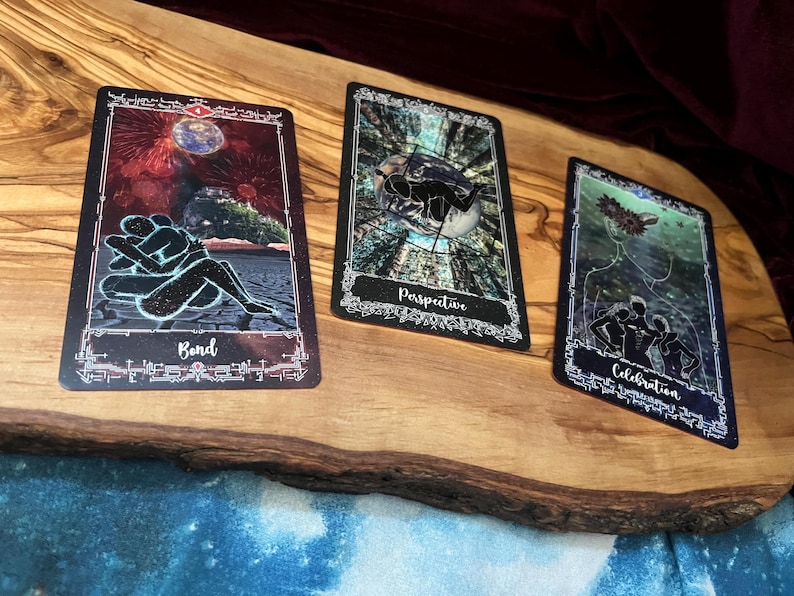 May include: Three rectangular tarot cards with black borders and intricate white filigree designs. The left card shows two figures embracing under a planet with the text Bond. The center card depicts a figure in a forest with the text Perspective. The right card shows three figures with the text Celebration.