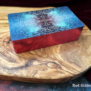 May include: A rectangular deck of cards featuring a blue and purple cosmic-themed back design with intricate white geometric patterns. The sides of the card deck are finished with a metallic red foil, creating distinct red gilded edges.